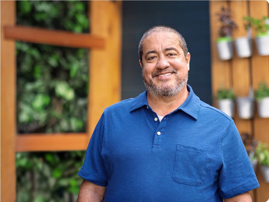 Man in a blue short-sleeved shirt stands outside surrounded by green plants