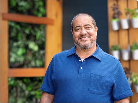 Man in a blue short-sleeved shirt stands outside surrounded by green plants