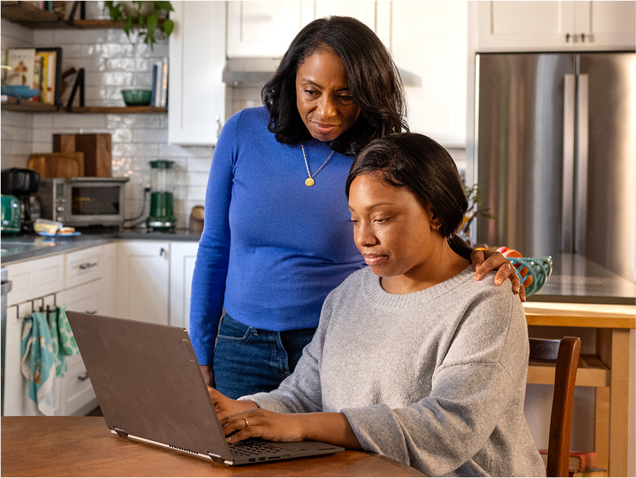 A woman at a kitchen table works on her laptop while another woman stands beside her, leaning in to see the computer’s screen