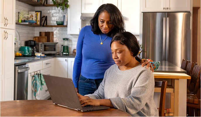 A woman at a kitchen table works on her laptop while another woman stands beside her, leaning in to see the computer’s screen